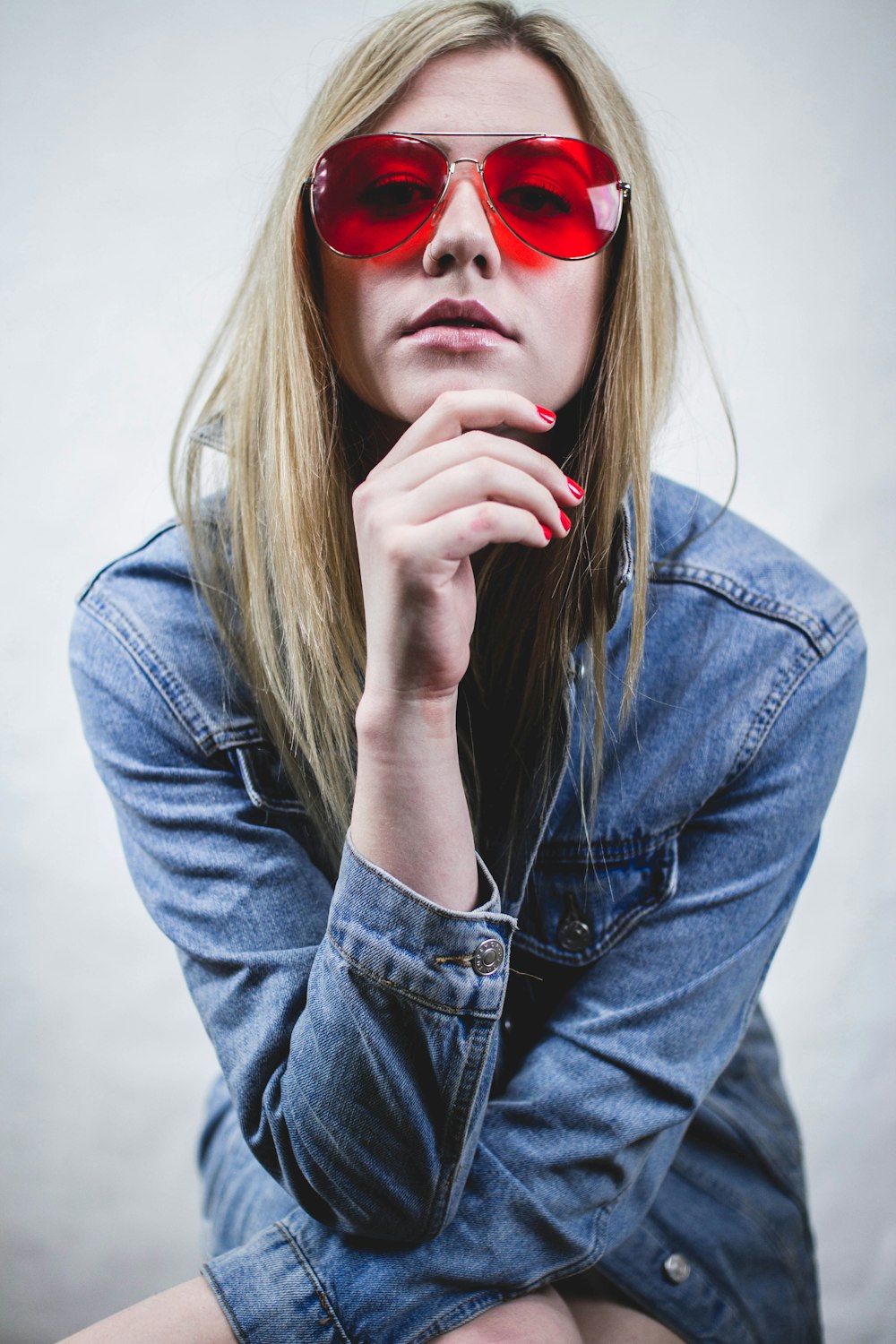 woman in blue denim jacket wearing red sunglasses and red manicure with hands on chin