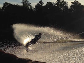 A person is waterskiing on a calm body of water, creating a striking arc of spray as they lean sharply to one side. The sunlight catches the spray, creating a shimmering effect against the silhouette of trees in the background.