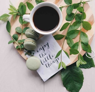 flat lay photography of leaves with cup of coffee and three macarons on chopping board