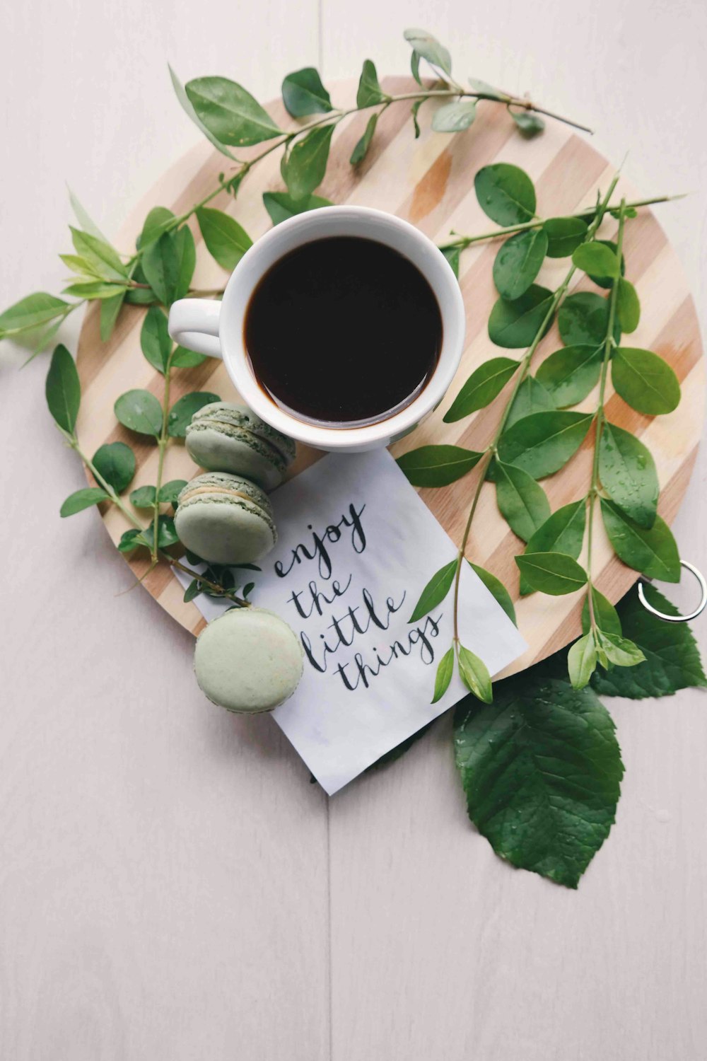 Photographie à plat de feuilles avec une tasse de café et trois macarons sur une planche à découper