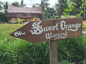 A rustic wooden signpost with the words 'Sweet Orange Warung' and '150m' directs towards a traditional thatched-roof building surrounded by green foliage and palm trees.