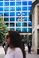 A modern office building with a blue reflective glass facade has the letters 'EDF' prominently displayed along with a logo. People are walking in the foreground; one person is on a phone, and others are blurred, suggesting movement. The setting is an urban environment with a combination of business and casual attire.