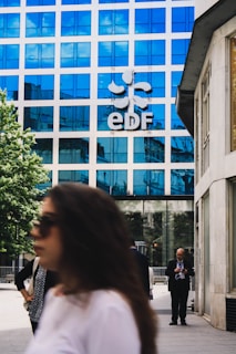 A modern office building with a blue reflective glass facade has the letters 'EDF' prominently displayed along with a logo. People are walking in the foreground; one person is on a phone, and others are blurred, suggesting movement. The setting is an urban environment with a combination of business and casual attire.
