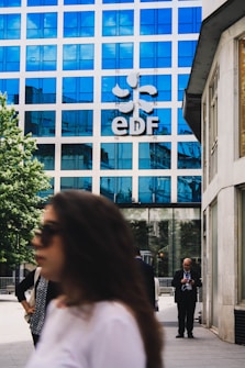 A modern office building with a blue reflective glass facade has the letters 'EDF' prominently displayed along with a logo. People are walking in the foreground; one person is on a phone, and others are blurred, suggesting movement. The setting is an urban environment with a combination of business and casual attire.