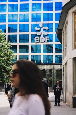 A modern office building with a blue reflective glass facade has the letters 'EDF' prominently displayed along with a logo. People are walking in the foreground; one person is on a phone, and others are blurred, suggesting movement. The setting is an urban environment with a combination of business and casual attire.