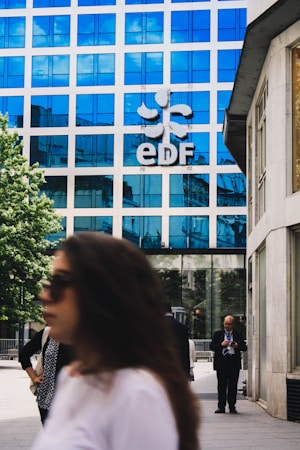 A modern office building with a blue reflective glass facade has the letters 'EDF' prominently displayed along with a logo. People are walking in the foreground; one person is on a phone, and others are blurred, suggesting movement. The setting is an urban environment with a combination of business and casual attire.
