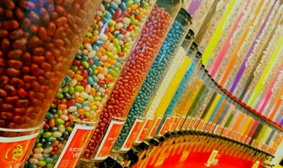 A vibrant display of large transparent canisters filled with various colored jelly beans, arranged in a neat line. The colors range from red, blue, yellow, green, orange, and pink, creating a rainbow effect. Each canister is labeled at the bottom, and the arrangement suggests a candy store or sweet shop setting.