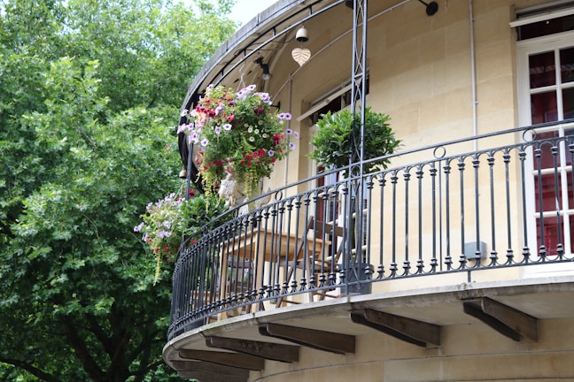 An elegant curved balcony adorned with hanging flower baskets and potted plants. The balcony features a black iron railing, with a wooden table and chairs placed near the wall of the building. Lush green trees provide a verdant backdrop, adding to the serene ambiance.