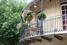 An elegant curved balcony adorned with hanging flower baskets and potted plants. The balcony features a black iron railing, with a wooden table and chairs placed near the wall of the building. Lush green trees provide a verdant backdrop, adding to the serene ambiance.