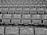 Rows of empty stadium seats, each with a numbered label attached to the top backrest, arranged in a symmetrical and repeating pattern.