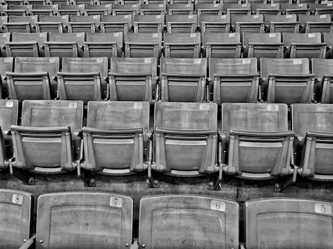 Rows of empty stadium seats, each with a numbered label attached to the top backrest, arranged in a symmetrical and repeating pattern.