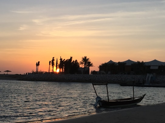 A tranquil sunset over a beachfront with silhouetted trees and a small boat on the water. The sky transitions from deep orange to light yellow, reflecting off the gentle waves. A row of tents or cabanas can be seen in the distance, adding a peaceful, vacation-like atmosphere.