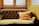 An elegant, vintage-style couch with a deep tufted backrest is positioned in front of large windows. The couch is upholstered in a muted gold fabric, and a matching, plush cushion with tassels is placed on it. The surrounding walls and window frames are painted in a warm, yellow hue, suggesting a cozy atmosphere.