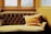 An elegant, vintage-style couch with a deep tufted backrest is positioned in front of large windows. The couch is upholstered in a muted gold fabric, and a matching, plush cushion with tassels is placed on it. The surrounding walls and window frames are painted in a warm, yellow hue, suggesting a cozy atmosphere.