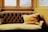 An elegant, vintage-style couch with a deep tufted backrest is positioned in front of large windows. The couch is upholstered in a muted gold fabric, and a matching, plush cushion with tassels is placed on it. The surrounding walls and window frames are painted in a warm, yellow hue, suggesting a cozy atmosphere.