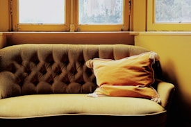 An elegant, vintage-style couch with a deep tufted backrest is positioned in front of large windows. The couch is upholstered in a muted gold fabric, and a matching, plush cushion with tassels is placed on it. The surrounding walls and window frames are painted in a warm, yellow hue, suggesting a cozy atmosphere.