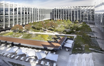 A modern architectural complex features a spacious courtyard with landscaped gardens filled with trees and greenery. Surrounding the courtyard, a large glass and metal building displays sleek, contemporary design with angular, geometric lines. The foreground contains an outdoor seating area with white umbrellas providing shade over tables and chairs.