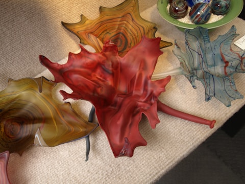 A group of colorful glass art pieces resembling leaves are arranged on a textured surface. The centerpiece is a striking red leaf with intricate swirls and patterns. Surrounding it are other leaves in varying shades of amber, blue, and green. In the background, there are spherical glass objects in a green bowl, featuring swirling patterns and vibrant colors.