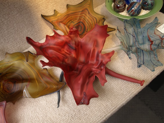 A group of colorful glass art pieces resembling leaves are arranged on a textured surface. The centerpiece is a striking red leaf with intricate swirls and patterns. Surrounding it are other leaves in varying shades of amber, blue, and green. In the background, there are spherical glass objects in a green bowl, featuring swirling patterns and vibrant colors.
