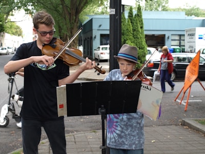 Two young individuals are playing violins on a city sidewalk. One is wearing sunglasses and a black shirt, the other is wearing a hat and tie-dye shirt. They are using a music stand, and a road sign is visible in the background. A few people are walking past.