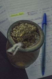 A traditional cup filled with yerba mate, accompanied by a metal straw. Next to it, a blue pen rests on open notebook pages that feature handwritten notes highlighted in neon yellow.