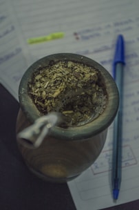 A traditional cup filled with yerba mate, accompanied by a metal straw. Next to it, a blue pen rests on open notebook pages that feature handwritten notes highlighted in neon yellow.