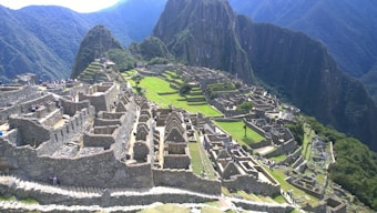 Ancient stone structures arranged on terraces with steep, green mountains in the background. The scene includes ruins of a historical site with grassy areas and remnants of buildings, showcasing intricate stonework. Some tourists are visible exploring the site, adding a sense of scale and activity.