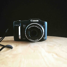 A digital camera sits on a wooden surface against a dark background. The camera is black with silver accents, featuring a 16x zoom lens and 16.0 megapixels.