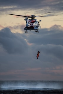 A helicopter hovers in the sky above a turbulent sea, with a rescue operation in progress. A person wearing bright orange rescue gear is suspended by a cable, being lifted from or lowered to the water below. The sky is cloudy with a dramatic, subdued sunset.