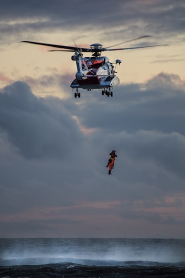 A helicopter hovers in the sky above a turbulent sea, with a rescue operation in progress. A person wearing bright orange rescue gear is suspended by a cable, being lifted from or lowered to the water below. The sky is cloudy with a dramatic, subdued sunset.