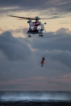A helicopter hovers in the sky above a turbulent sea, with a rescue operation in progress. A person wearing bright orange rescue gear is suspended by a cable, being lifted from or lowered to the water below. The sky is cloudy with a dramatic, subdued sunset.