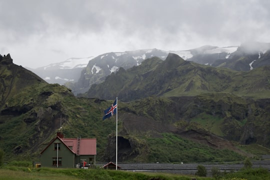 white concrete house with white, blue, and red cross flag near mountain range covered in green vegetation at daytime in Thórsmörk Iceland