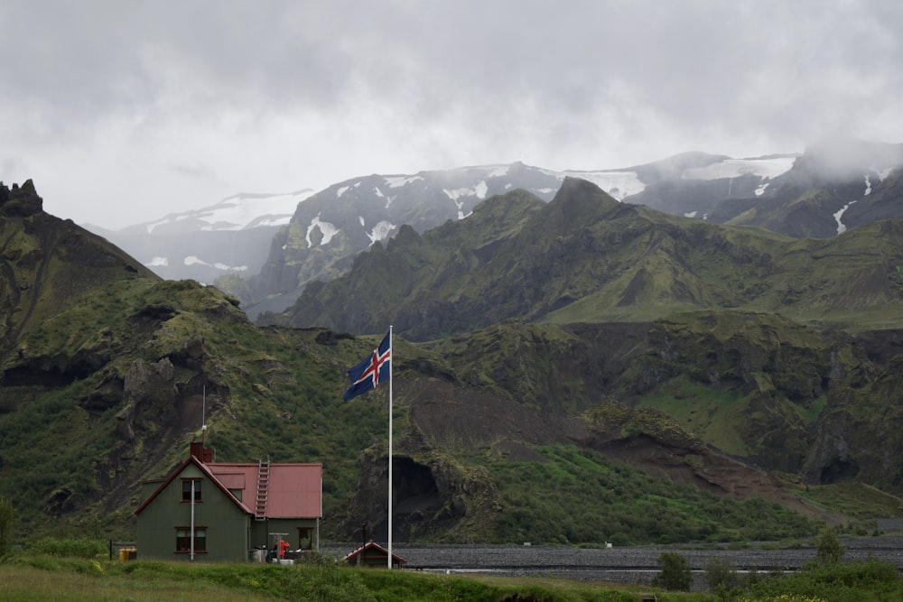 white concrete house with white, blue, and red cross flag near mountain range covered in green vegetation at daytime
