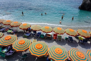 Brightly colored orange and green striped beach umbrellas are lined up on a pebbled shore. People are relaxing under the umbrellas on sun loungers while others are swimming and playing in the clear blue sea. The scene conveys a lively and summery atmosphere.