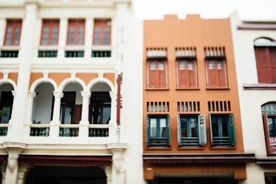 Two adjacent buildings with architectural design featuring multiple windows and arches. The left building has a classic colonial style with white columns and green shutters, while the right building is more modern with a terracotta facade and red shutters.