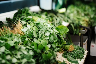 A variety of fresh leafy greens are piled on a table, including spinach and other types of herbs. The greens are vibrant and appear freshly harvested, with some roots still attached. The setting suggests an environment like a market or grocery store.