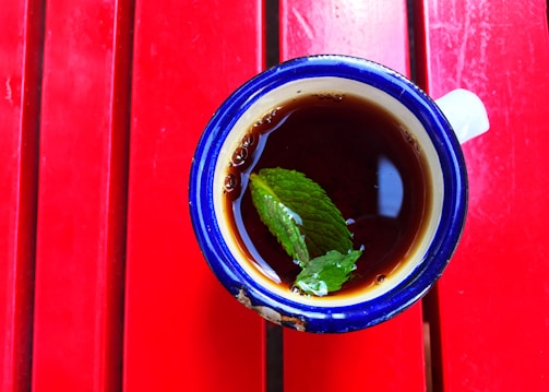 A cup with a dark beverage, garnished with a fresh green mint leaf, sits against a bright red wooden surface. The cup has a blue rim and appears to be ceramic. The red surface provides a striking contrast to the deep color of the beverage and the mint leaf.