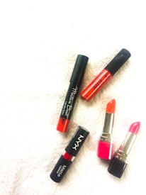 Five lipsticks are arranged on a textured white surface. The lipsticks include a mix of matte and glossy finishes with two having a traditional lipstick shape while the rest are in tube form. The colors range from orange to pink to red, with black caps and metallic accents.