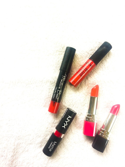 Five lipsticks are arranged on a textured white surface. The lipsticks include a mix of matte and glossy finishes with two having a traditional lipstick shape while the rest are in tube form. The colors range from orange to pink to red, with black caps and metallic accents.