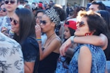 A group of young people gathered outdoors, some wearing sunglasses and colorful clothing. There are several women and men interacting closely, with one woman wearing a headscarf and another sporting rainbow wristbands. The atmosphere appears lively and social.