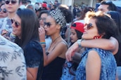 A group of young people gathered outdoors, some wearing sunglasses and colorful clothing. There are several women and men interacting closely, with one woman wearing a headscarf and another sporting rainbow wristbands. The atmosphere appears lively and social.