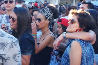 A group of young people gathered outdoors, some wearing sunglasses and colorful clothing. There are several women and men interacting closely, with one woman wearing a headscarf and another sporting rainbow wristbands. The atmosphere appears lively and social.