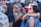 A group of young people gathered outdoors, some wearing sunglasses and colorful clothing. There are several women and men interacting closely, with one woman wearing a headscarf and another sporting rainbow wristbands. The atmosphere appears lively and social.