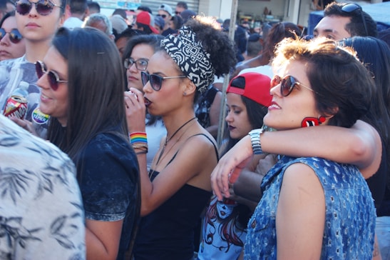 A group of young people gathered outdoors, some wearing sunglasses and colorful clothing. There are several women and men interacting closely, with one woman wearing a headscarf and another sporting rainbow wristbands. The atmosphere appears lively and social.