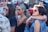 A group of young people gathered outdoors, some wearing sunglasses and colorful clothing. There are several women and men interacting closely, with one woman wearing a headscarf and another sporting rainbow wristbands. The atmosphere appears lively and social.