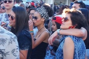 A group of young people gathered outdoors, some wearing sunglasses and colorful clothing. There are several women and men interacting closely, with one woman wearing a headscarf and another sporting rainbow wristbands. The atmosphere appears lively and social.