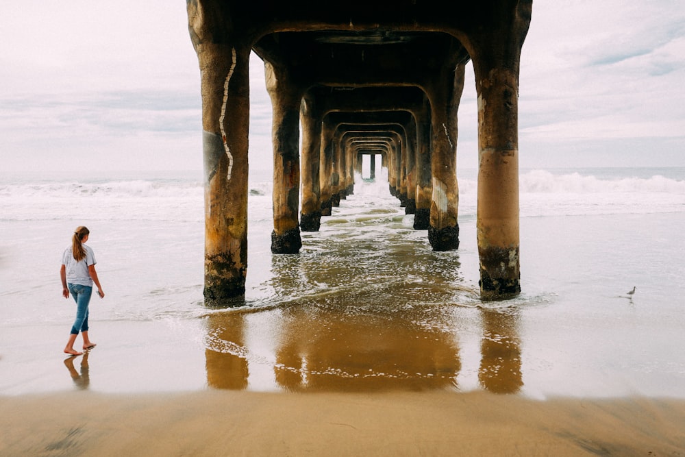 woman walking on seashore towards bridge pillars