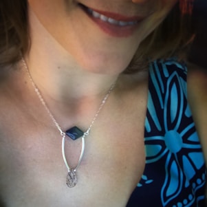 A close-up view of a woman wearing a necklace with a geometric pendant and a patterned blue dress. The necklace features a delicate chain and two distinct pendants with unique shapes.