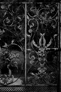 An intricately designed wrought iron gate with various floral and vine patterns. The iron appears to be aged and slightly weathered, with a dark, monochromatic color scheme. In the background, there are blurred, out-of-focus trees and foliage, suggesting an outdoor setting.