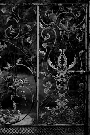 An intricately designed wrought iron gate with various floral and vine patterns. The iron appears to be aged and slightly weathered, with a dark, monochromatic color scheme. In the background, there are blurred, out-of-focus trees and foliage, suggesting an outdoor setting.